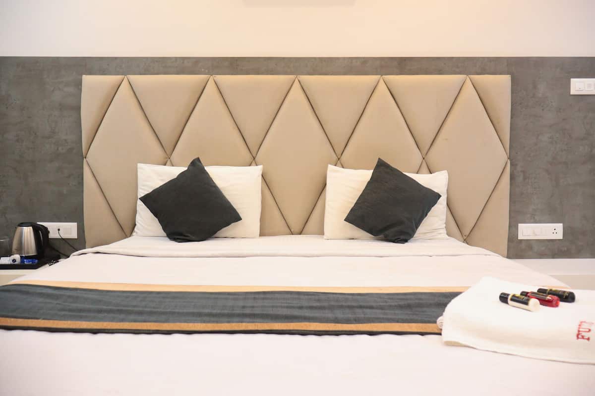 Habitación de hotel con una cama grande, dos almohadas, una mesita de noche con tetera y una colcha.