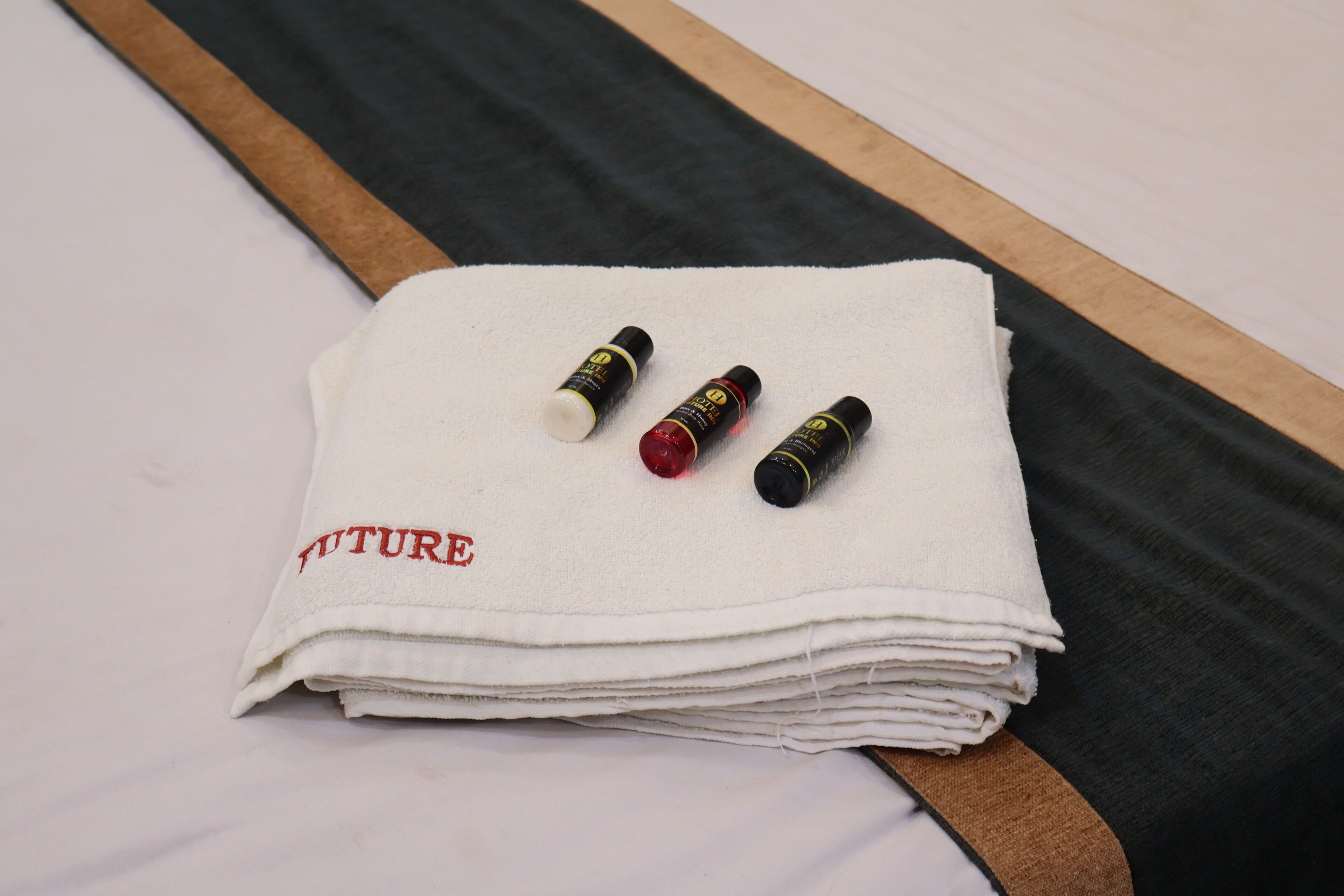 A neatly folded white towel with the word "FUTURE" embroidered on it, placed on a bed with a wooden headboard and a dark-colored blanket.