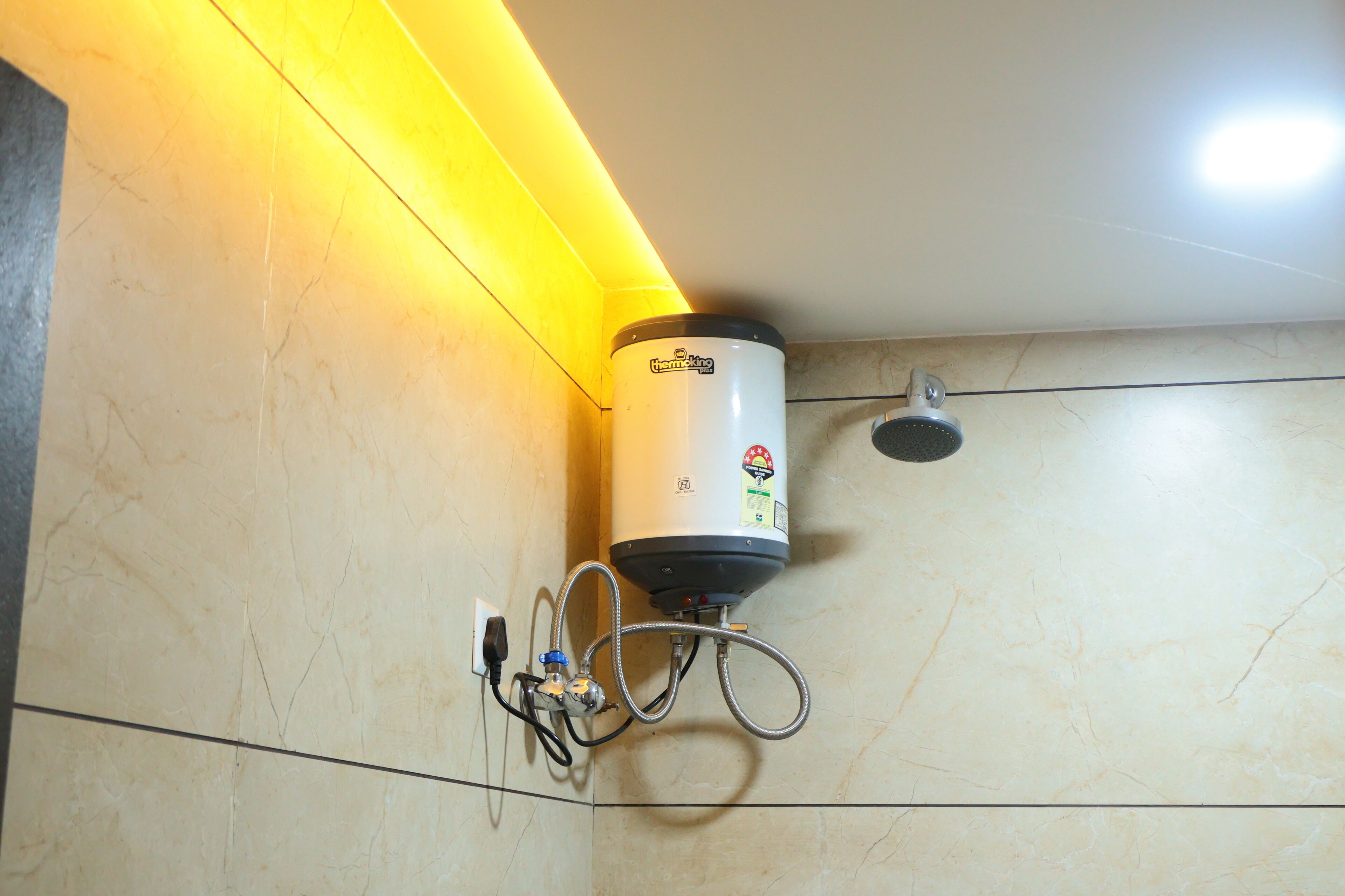 Um aquecedor de água fixado em uma parede revestida com azulejos, com uma tomada elétrica e válvulas de controle visíveis.