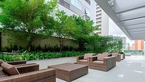 An outdoor seating area with wicker furniture and a tiled floor, surrounded by greenery and a building facade. - Studio Central no Prédio 7Th Avenue, com ar e vaga (Curitiba)