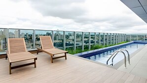 A rooftop pool area with two wooden lounge chairs and a view of the cityscape. - Studio Central no Prédio 7Th Avenue, com ar e vaga (Curitiba)