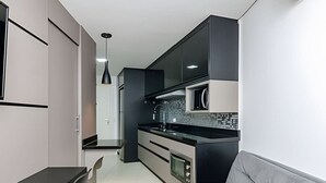 A modern kitchen with a black countertop, a built-in oven, and a grey sofa in the living area. - Studio Central no Prédio 7Th Avenue, com ar e vaga (Curitiba)