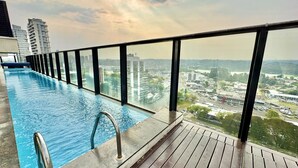 A rooftop infinity pool with a wooden deck and glass railings, overlooking a cityscape with buildings and a river.