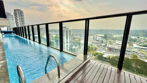 Una piscina infinitum al terrat, amb terrassa de fusta i barana de vidre, amb vista a un paisatge urbà amb edificis i un riu.