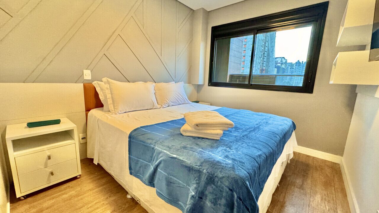 A hotel room with a bed, a nightstand, a window with a city view, and a wall-mounted shelf.