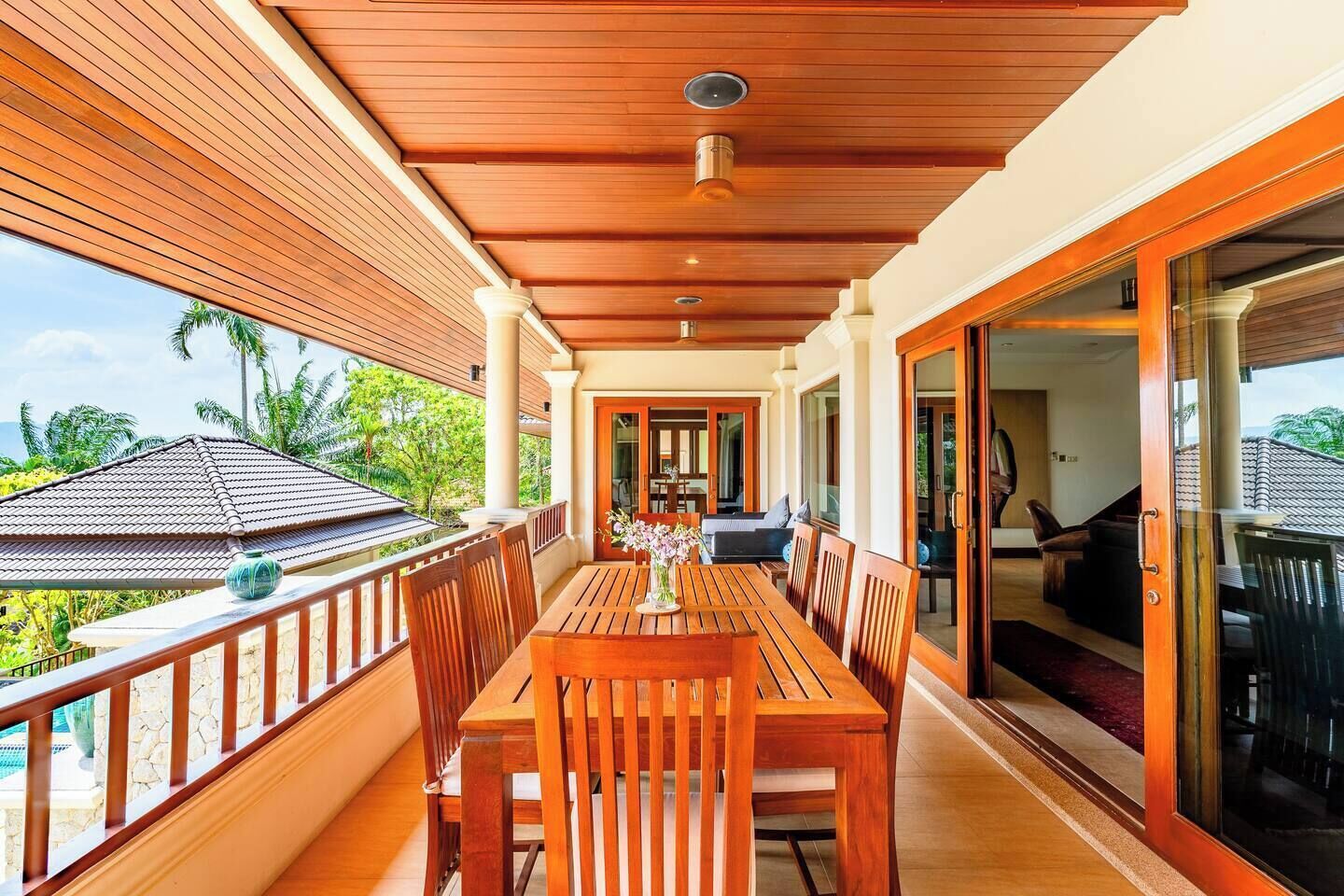 Una terraza de madera con una mesa y sillas, un vidrio que da a una habitación y vistas a un tejado con tejas y vegetación.