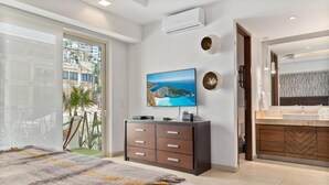 Ein modernes Schlafzimmer mit einem großen Bett, einem an der Wand montierten Fernseher, einer Glastür zu einem Balkon sowie einem Badezimmer mit Waschtisch und Spiegel.