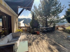 En træterrasse med udendørs møbler, udsigt til bjerge og en bygning med en overdækket terrasse.