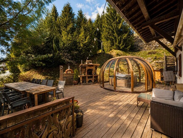 An outdoor deck with a wooden table and chairs, a glass-enclosed seating area, and a brick fireplace.
