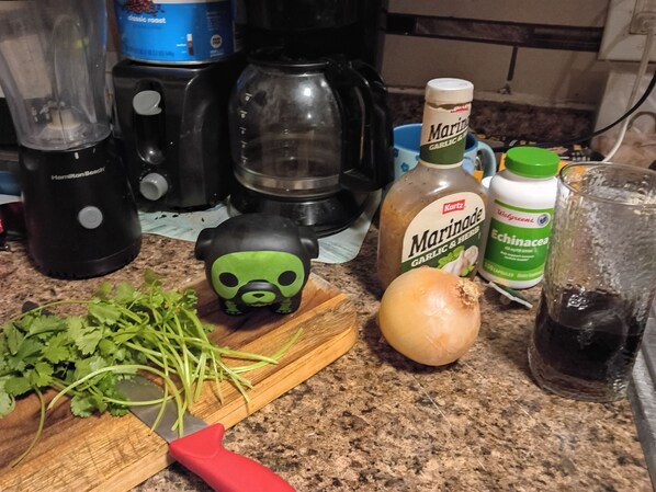 Una encimera de cocina con una licuadora, una cafetera, una botella de adobo, una botella de equinácea, un vaso y una tabla de cortar con un cuchillo y hierbas picadas.