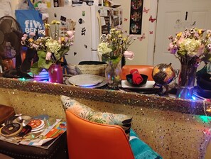 Ein Zimmer mit einer dekorativen Ablage, verschiedenen Vasen mit Blumen, einem Ventilator und einem Sessel mit Kissen.