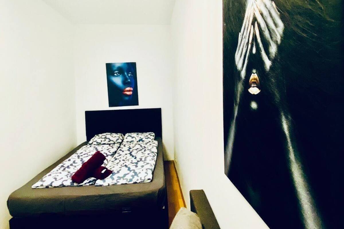 Quarto pequeno com uma cama de solteiro, cabeceira preta, colcha estampada e travesseiro vermelho. Há um quadro na parede e um grande quadro abstrato à direita.