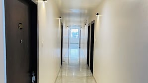 Corredor de hotel com uma porta escura à esquerda, uma placa de número e vista para o hall de entrada ao fundo.