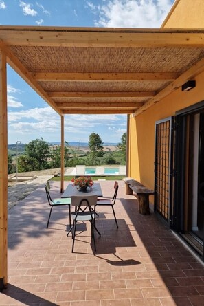 A covered patio with a table and chairs, a bench, and a view of a swimming pool and landscape.