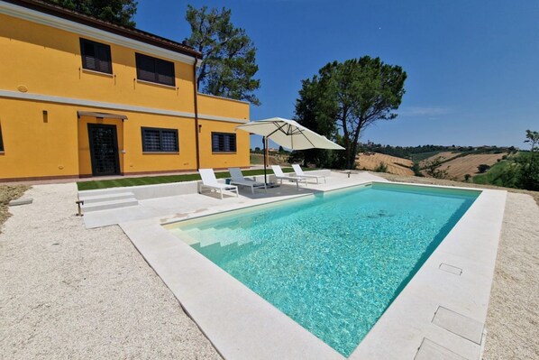 A swimming pool with white lounge chairs and an umbrella, surrounded by a yellow building and trees.