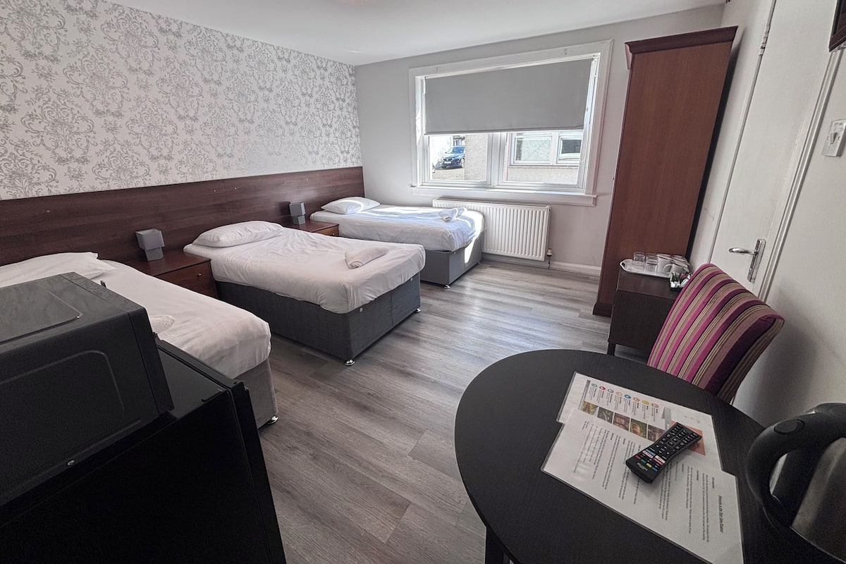 A hotel room with two beds, a small table with a remote control, and a window with a view of the outside.