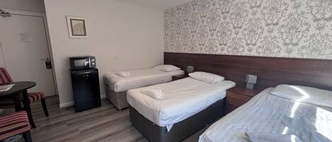 A hotel room with two single beds, a small table with a chair, a desk, a trash bin, and a wall with a patterned wallpaper.