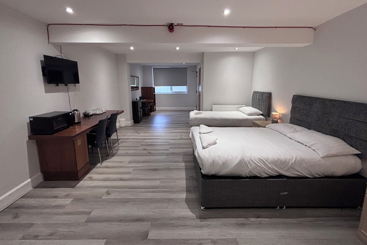 A modern hotel room with a large bed, a desk with a computer, a flat-screen TV, and a view of the hallway.