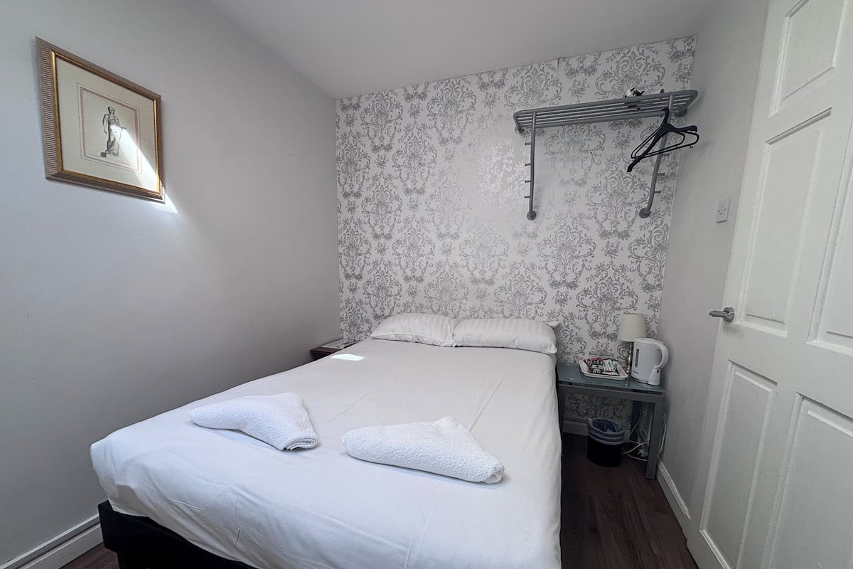 A neatly made bed with two towels, a bedside table with a lamp and a kettle, and a wall-mounted drying rack.