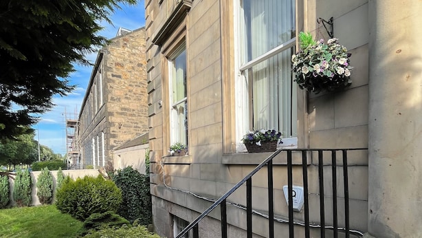 A stone building with a staircase leading to the entrance, surrounded by greenery and a clear sky.