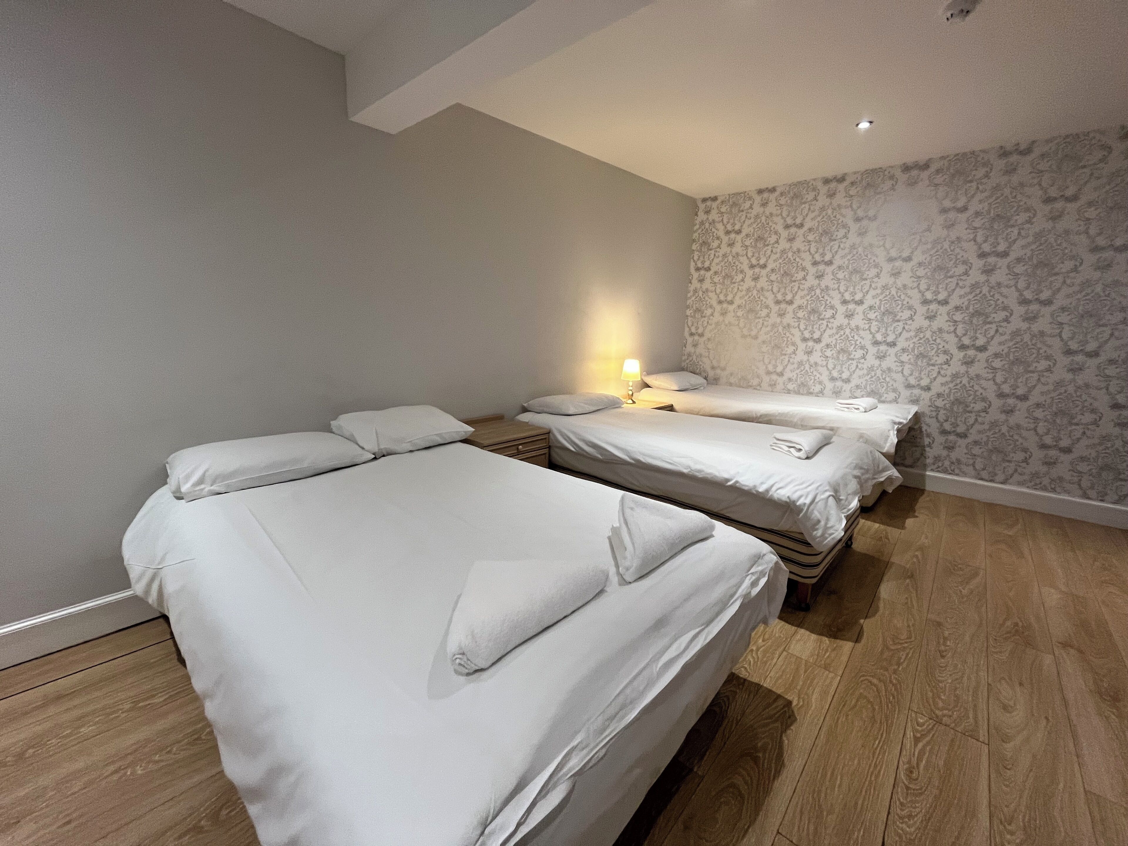 A hotel room with two single beds, a wooden floor, a patterned wallpaper on one wall, and a nightstand with a lamp.