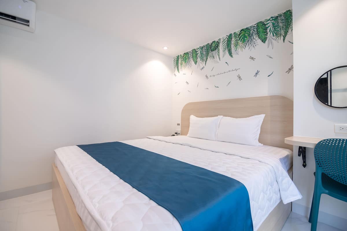 A neatly made bed with white and blue bedding, a wooden headboard, a wall-mounted air conditioner, and a wall decoration with green leaves and birds.