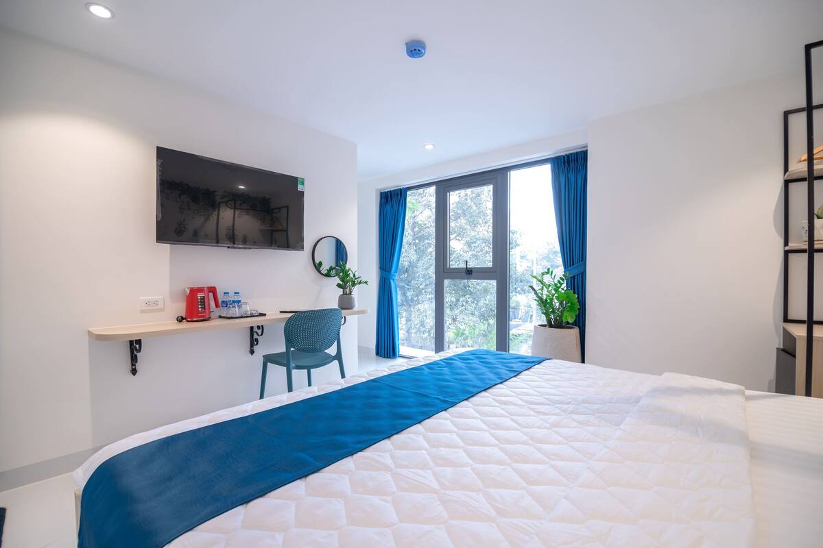 A hotel room with a large bed, a desk with a chair, a TV mounted on the wall, and a window with curtains.