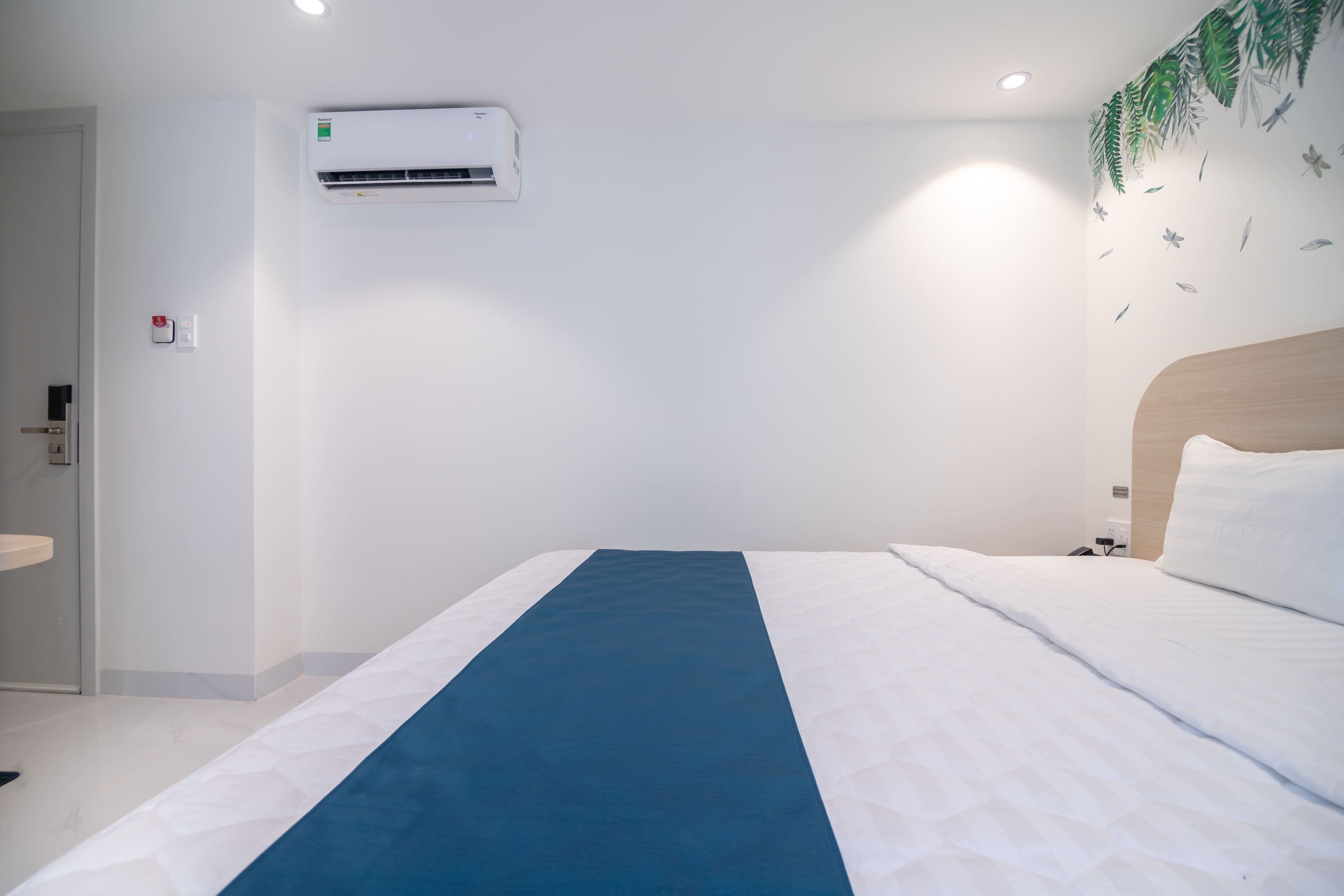 Una habitación de hotel con cama, aire acondicionado instalado en la pared, cabecero de madera y un adhesivo mural con hojas y pájaros.