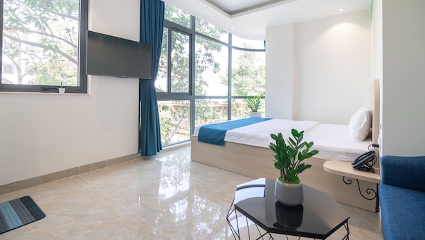 A modern hotel room with a large bed, a small table with a plant, a blue sofa, and a view of trees through large windows. - Bin Bin Hotel 15 - Near Feliz En Vista (Ho Chi Minh City)