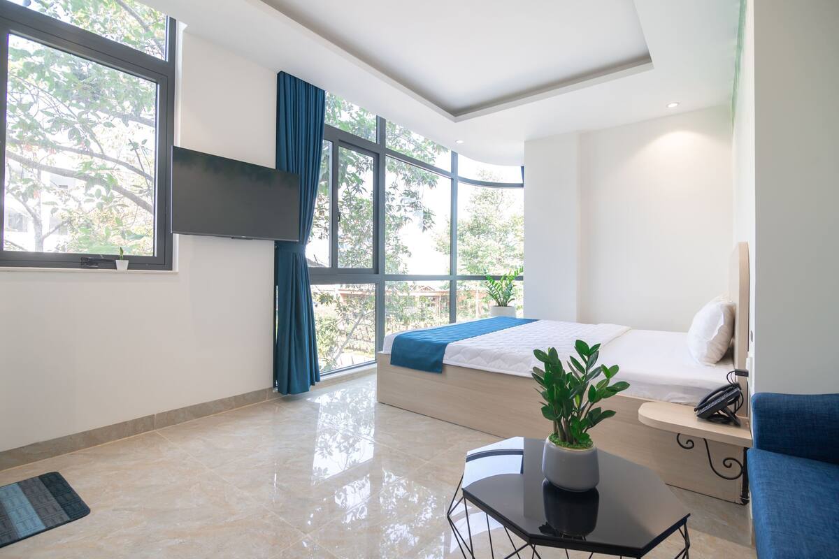 A modern hotel room with a large bed, a small table with a plant, a blue sofa, and a view of trees through large windows.