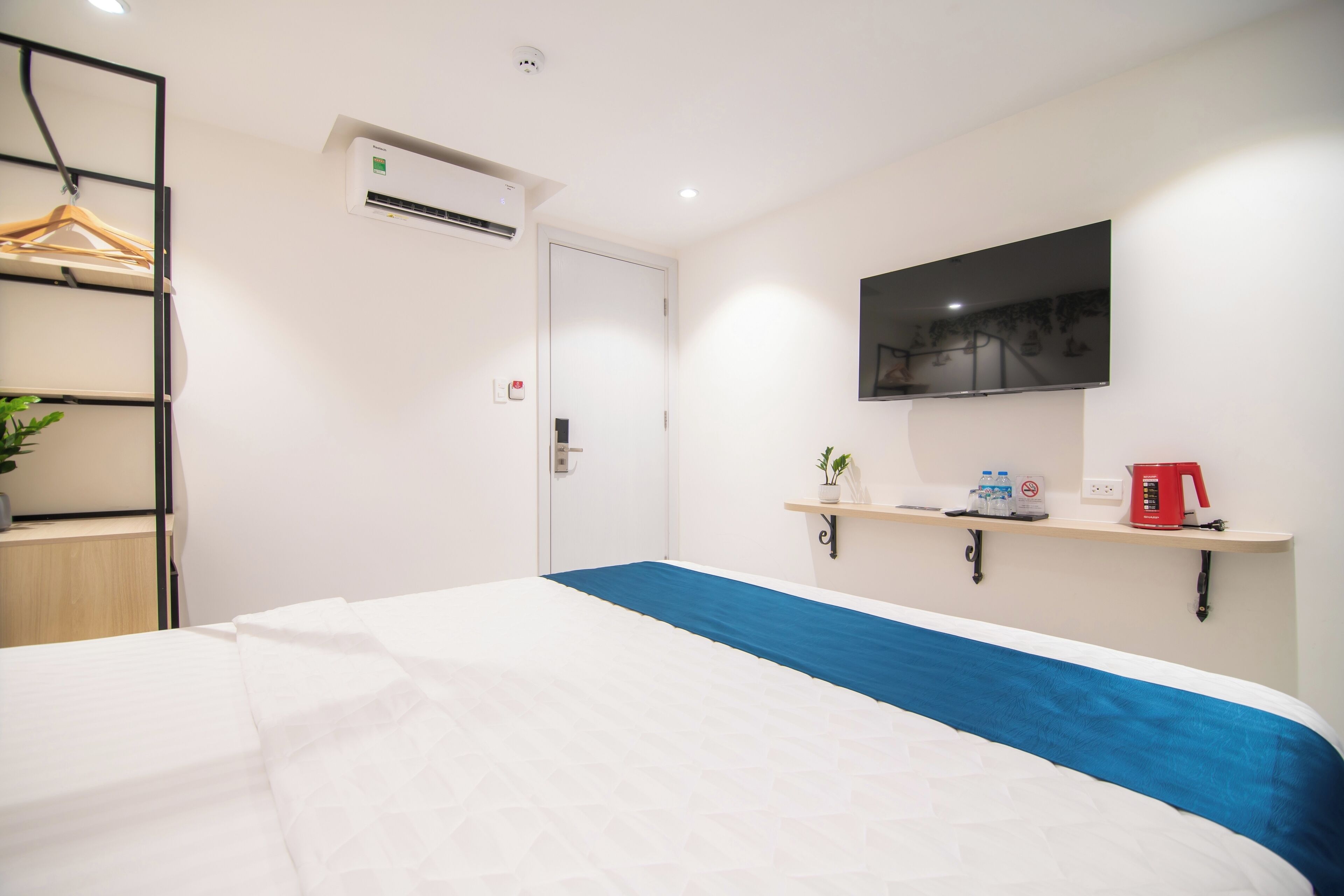 Una habitación de hotel con una cama, un televisor montado en la pared, un estante con una planta y una tetera, y un equipo de aire acondicionado.