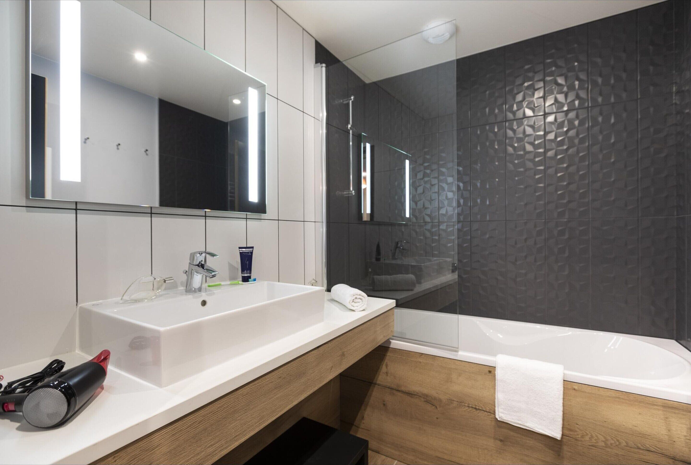 Une salle de bain moderne avec un grand miroir, un lavabo blanc, une baignoire et un meuble-lavabo en bois.