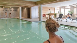 An indoor swimming pool with a clear view of the pool's edge, a wooden wall with a sign, and a seating area with lounge chairs and a person relaxing. - Apartment in France With Spa and Pool (Les Belleville)