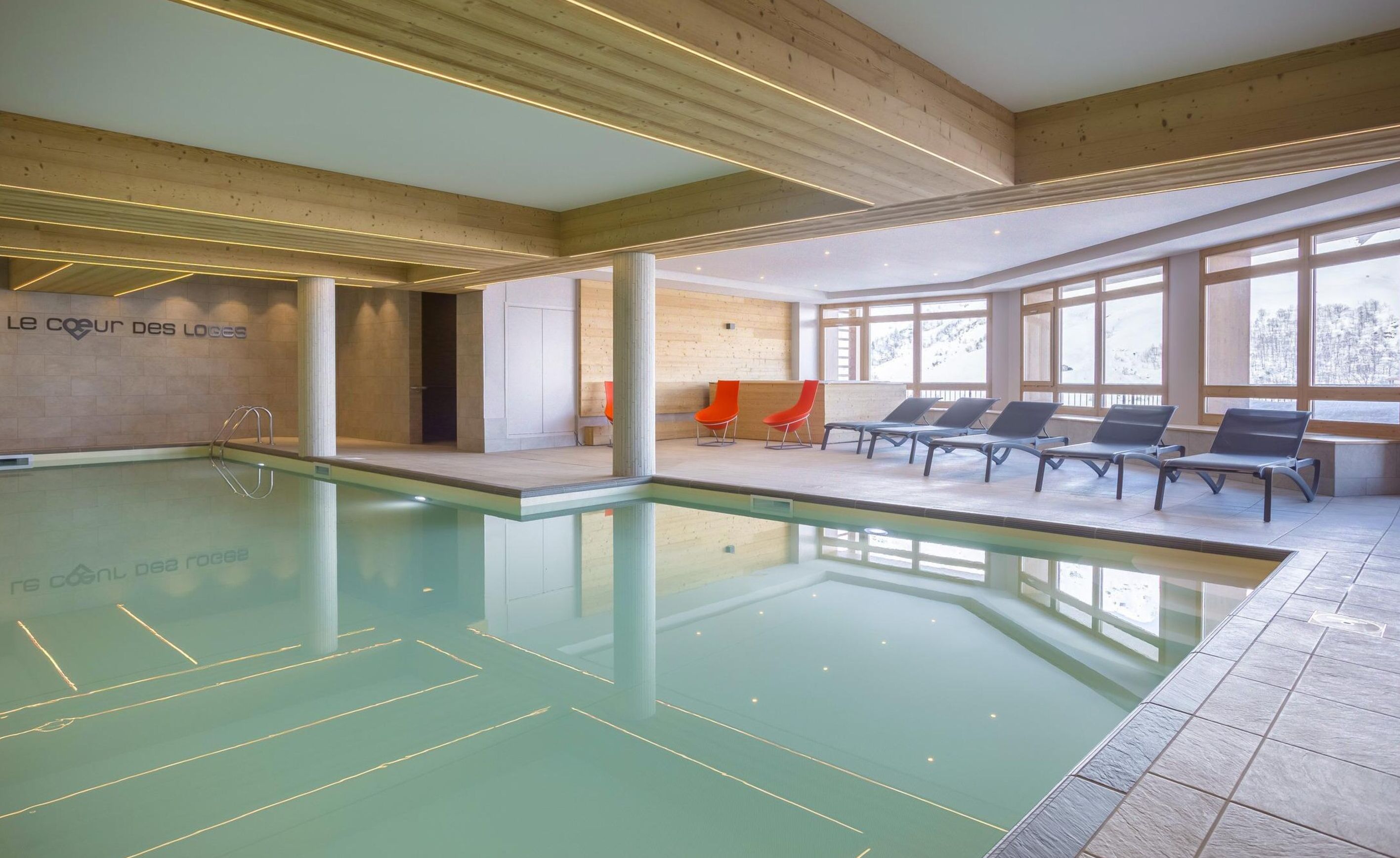 Une piscine intérieure moderne avec des couloirs bien délimités, un espace détente avec des fauteuils et de grandes fenêtres laissant entrer la lumière naturelle.