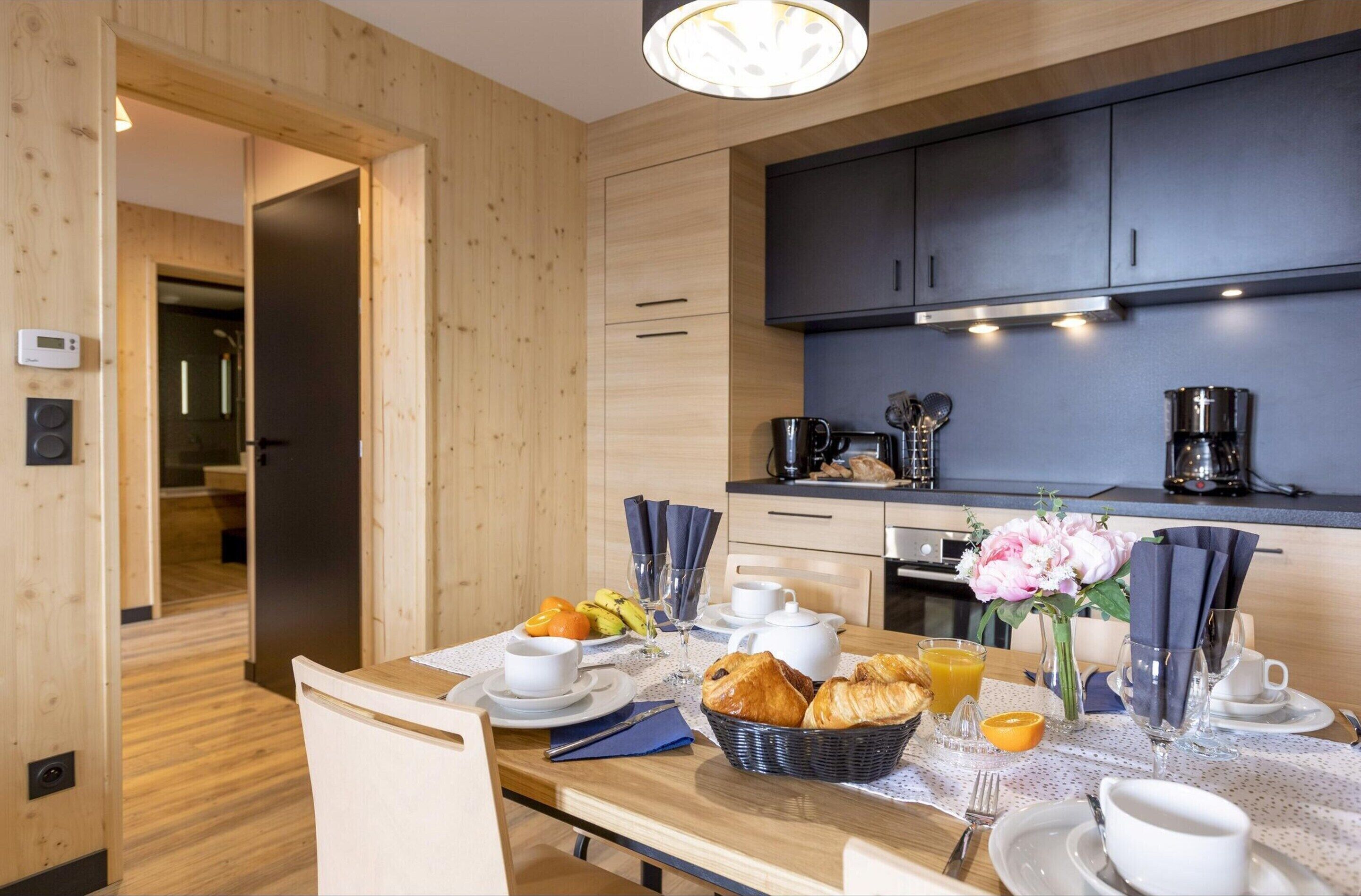 Cocina moderna con muebles de madera y una mesa para el desayuno con cruasanes, jugo de naranja y un jarrón de flores.