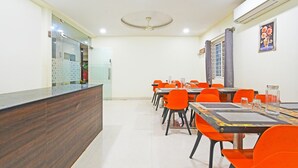 Una habitación de hotel con una recepción, un área de comedor con sillas y mesas anaranjadas, un ventilador de techo y un equipo de aire acondicionado.