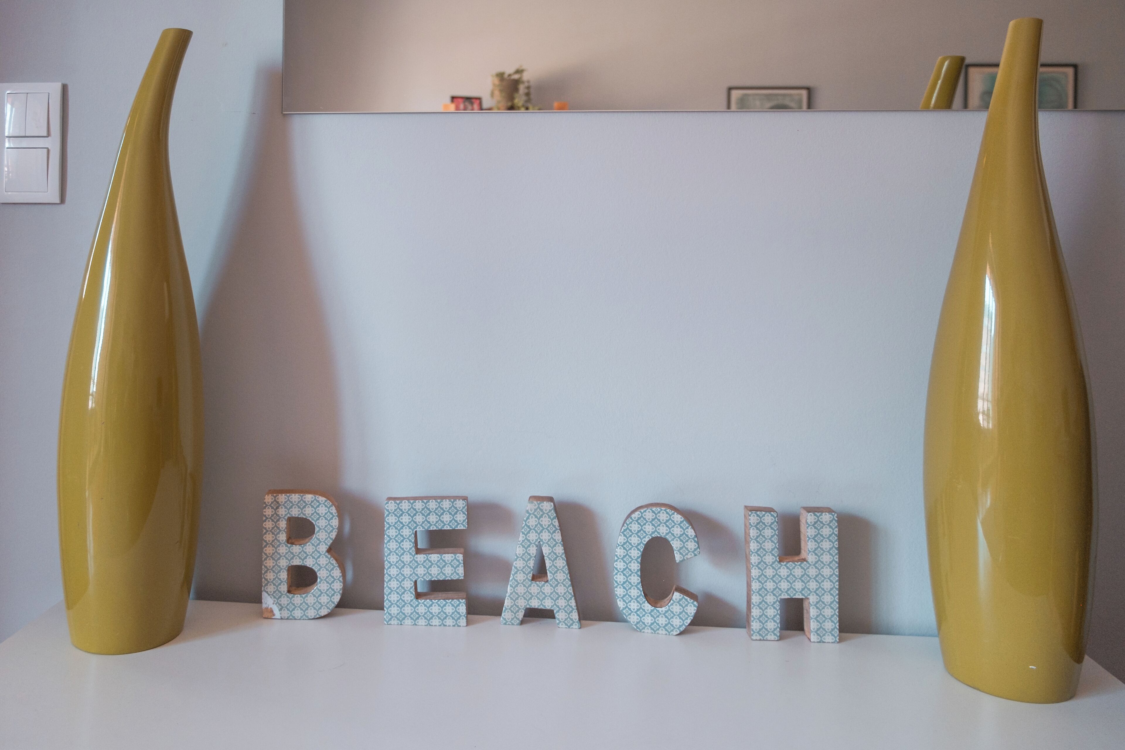 Eine dekorative Anordnung mit dem Wort "BEACH" in der Mitte, flankiert von je einer hohen Vase auf beiden Seiten.