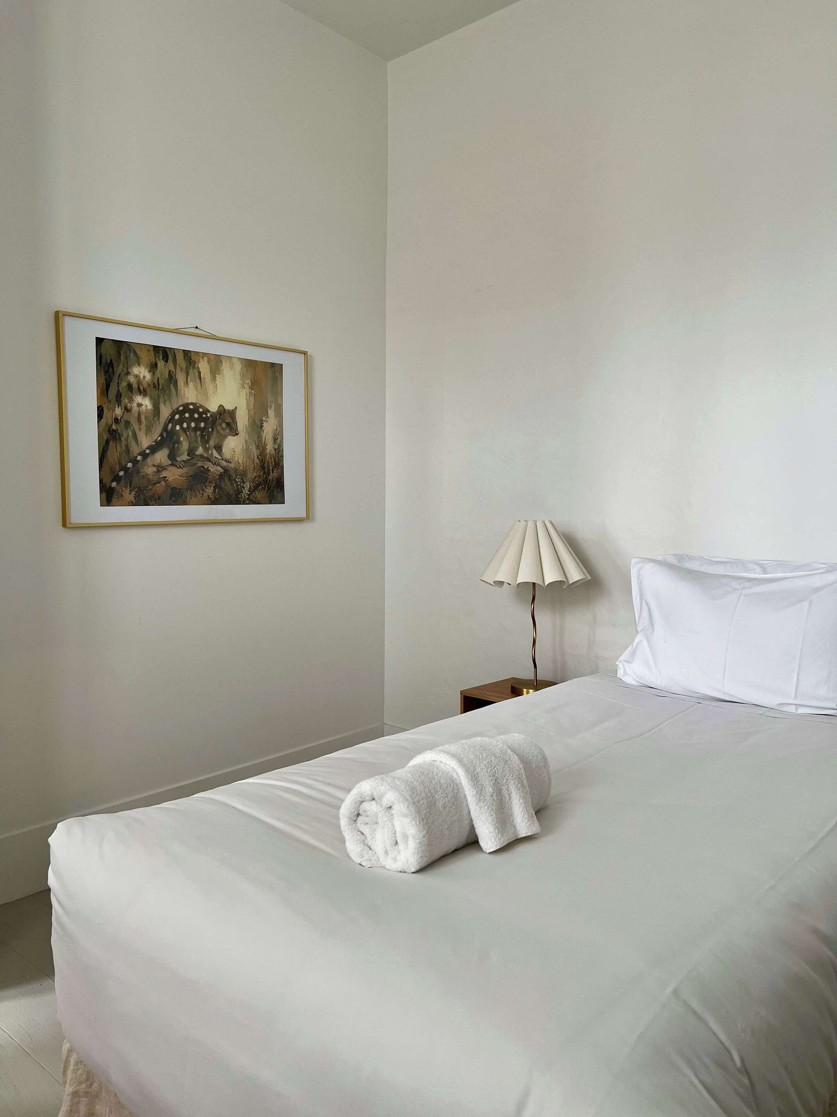 Een hotelkamer met een net opgemaakt bed, een wit kussen, een opgerolde witte handdoek, een lamp en een ingelijst schilderij aan de muur.