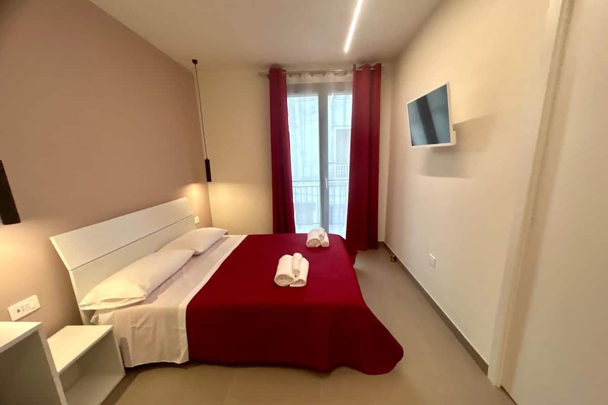 A hotel room with a bed, a TV on the wall, and a window with curtains.