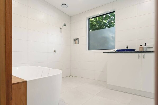 A modern bathroom with white tiles, a bathtub, a shower, and a window with a view of greenery. - Hidden Retreat - Off Grid (West Launceston)