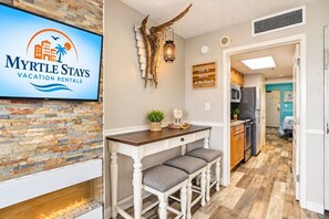 A modern kitchen with a built-in stove, a dining area with a wooden table and bar stools, and a sign for Myrtle Stays Vacation Rentals. - Oceanfront Retreat with Resort Pools (Myrtle Beach)