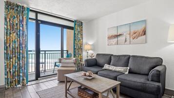 Ein Wohnzimmer mit grauem Sofa, einem Holztisch und Blick auf den Strand durch eine Glastür.