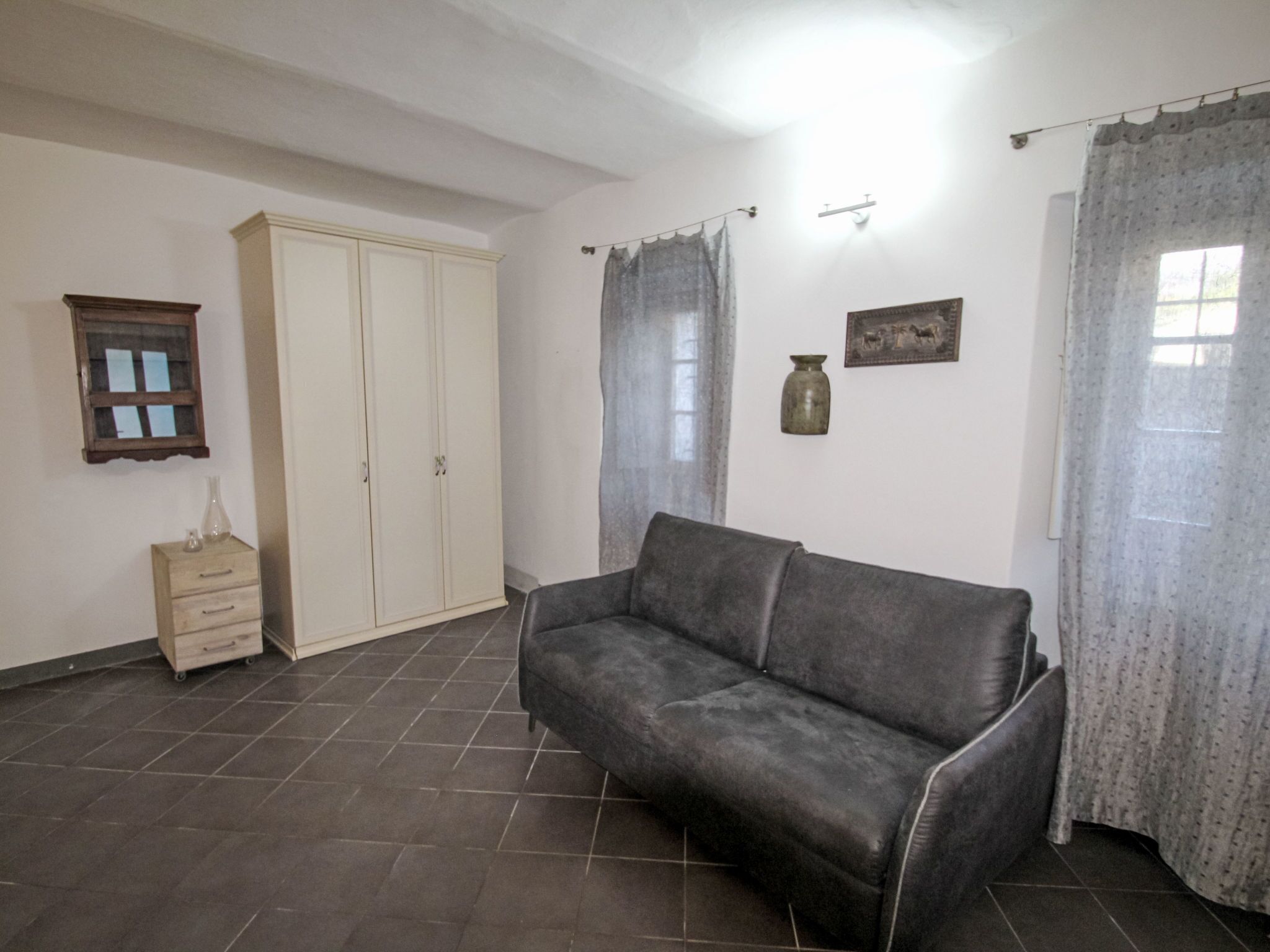 Una sala de estar con un sofá gris, un armario de madera, una mesita con un jarrón y una ventana con cortinas.