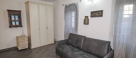 Una sala de estar con un sofá gris, un armario de madera, una mesita con un jarrón y una ventana con cortinas.