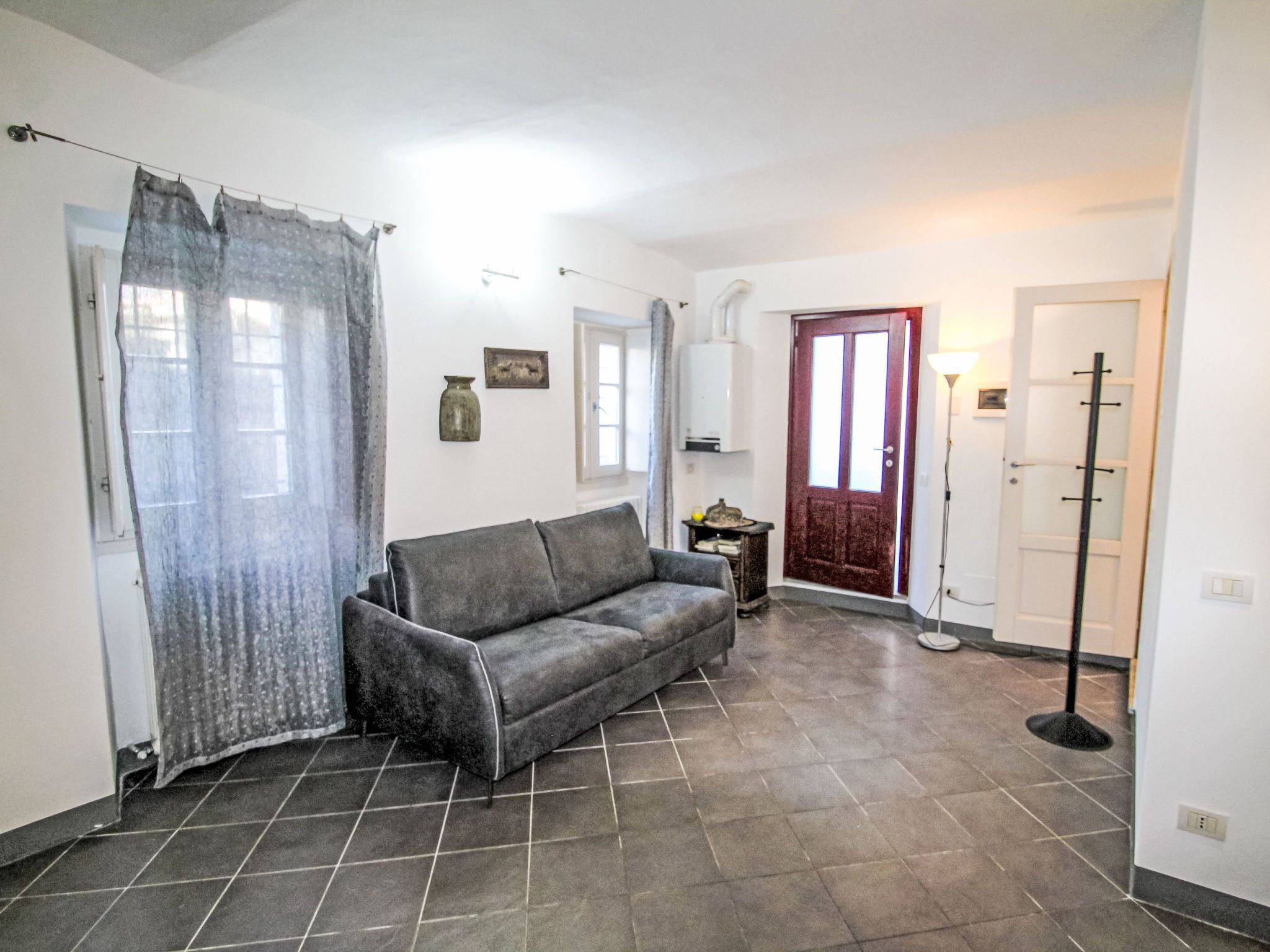 Ein Wohnzimmer mit grauem Sofa, rotem Tür und gefliestem Boden.