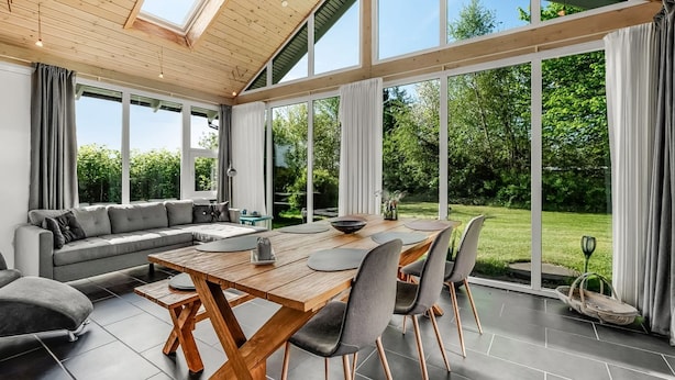 Ein modernes Interieur mit Holzbalkendecke, großen Fenstern, einer Essecke mit Tisch und Stühlen sowie einem grauen Sofa.
