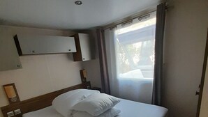A hotel room with a bed, a nightstand, a wall-mounted shelf, a window with curtains, and a door. - Ideal rental campsite oasis village 5⭐️ (Puget-sur-Argens)