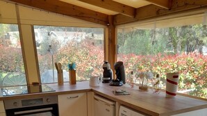 A compact kitchen with a built-in oven, dishwasher, and washing machine. It features wooden beams, large windows, and a view of a garden with red foliage. - Ideal rental campsite oasis village 5⭐️ (Puget-sur-Argens)