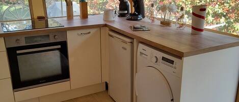 Een compacte keuken met een ingebouwde oven, vaatwasser en wasmachine. Houten balken, grote ramen en uitzicht op een tuin met rood herfstblad.