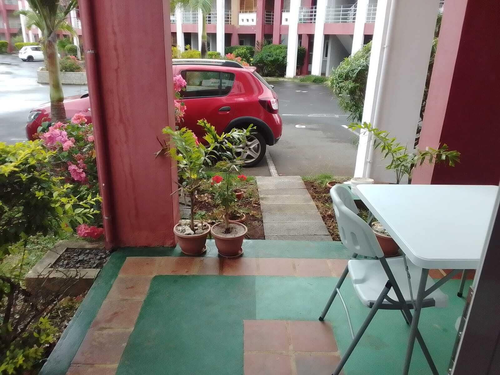 Ein überdachter Außenbereich mit einem roten Auto auf der Straße, Blumentöpfen und einem weißen Tisch mit einem Stuhl.